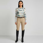 Woman wearing a patterned top, beige riding pants, and black riding boots on a gray background